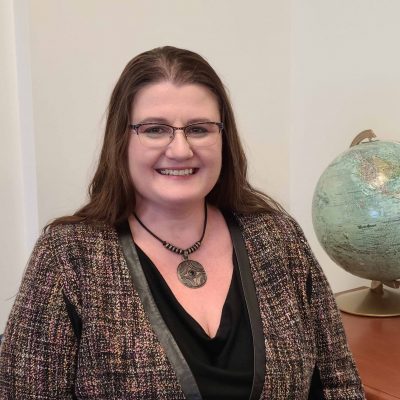 Jamie Baker Woman with glasses smiling, wearing a checkered blazer and necklace, with a globe in the background.