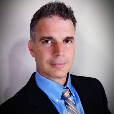 Male with short brown hair wearing a blue button shirt orange, white and blue striped tie and sports coat. 
